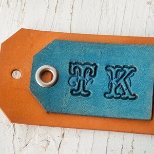 Pode incluir: Uma etiqueta de couro com um inserto de couro azul. O inserto tem um ilh&oacute; prateado e as letras "FK" em relevo em uma fonte decorativa.