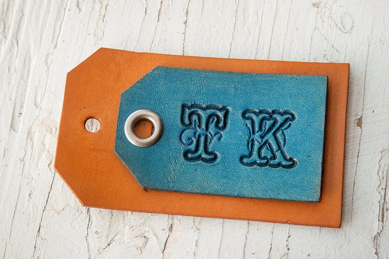 Pode incluir: Uma etiqueta de couro com um inserto de couro azul. O inserto tem um ilh&oacute; prateado e as letras "FK" em relevo em uma fonte decorativa.