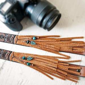 May include: Two brown leather camera straps with fringe and turquoise beads. The straps have a decorative design and are attached to black metal clips.