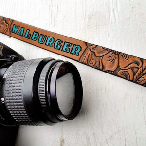 May include: A brown leather camera strap with an embossed floral design and the text "WALBURGER" in turquoise blue lettering.