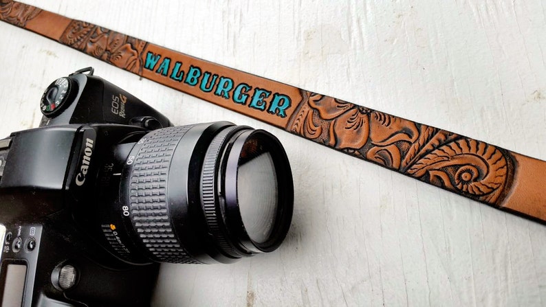 May include: A brown leather camera strap with an embossed floral design and the text "WALBURGER" in turquoise blue lettering.