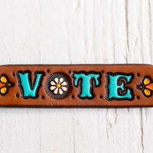 May include: A brown leather accessory with the word "VOTE" in turquoise letters. The design includes a daisy and floral accents in orange and yellow. The item is set against a white, textured background.