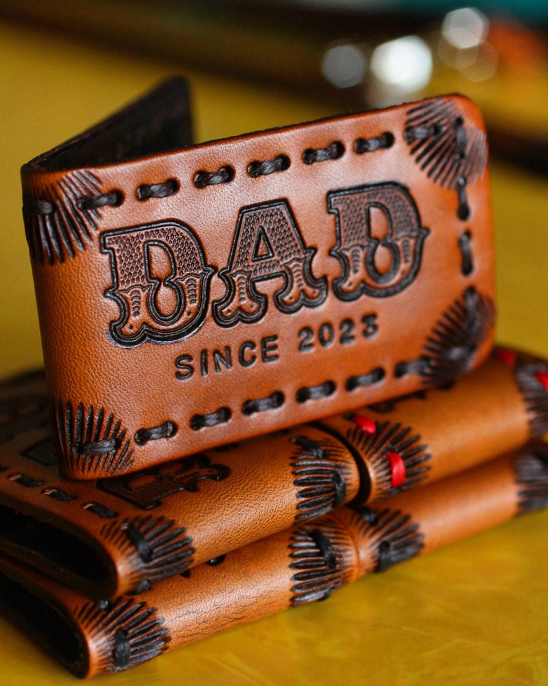 May include: A brown leather wallet with black stitching and a decorative design. The wallet has the word 'DAD' embossed on the front, along with the text 'SINCE 2023'.