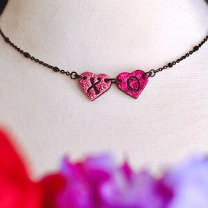 May include: A delicate black chain necklace with two pink glitter heart pendants. The hearts have the letters "X" and "O" printed on them.