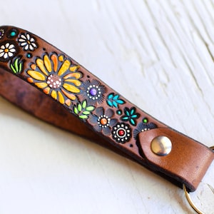 May include: A brown leather keychain with a floral design. The flowers are painted in yellow, orange, green, blue, and purple. The keychain has a silver metal ring.