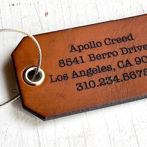 Könnte beinhalten: Ein brauner Leder-Gepäckanhänger mit dem Namen "Apollo Creed" und Adressinformationen, die in Schwarz eingraviert sind. Der Anhänger ist an einer silberfarbenen Metallschlaufe befestigt.