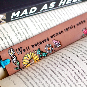 May include: A brown leather bookmark featuring the quote "Well behaved women rarely make history." It's adorned with colourful floral and butterfly designs and a blue tassel, resting on a book.