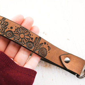 Könnte beinhalten: Ein brauner Leder-Schlüsselanhänger mit einem schwarzen, gravierten Design aus Kakteen, Blumen und Sternen. Der Schlüsselanhänger hat einen silbernen Schlüsselring aus Metall.