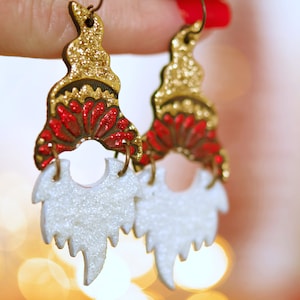 May include: A pair of gold and red glitter earrings with white glitter beard accents. The earrings are shaped like a gnome's hat and beard.