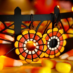 May include: A pair of orange, yellow and white ceramic earrings with a geometric design. The earrings are hanging from a black metal rack with a spiderweb design. The earrings are surrounded by candy corn.