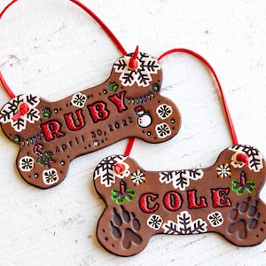 Könnte beinhalten: Zwei braune, knochenförmige Hundeornamente mit roter Schrift und Akzenten. Eines trägt die Aufschrift "RUBY April 30, 2022" und das andere "COLE". Beide haben Schneeflocken- und Pfotenabdruck-Designs mit roten Aufhängeschnüren.