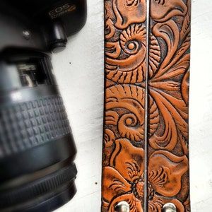 May include: A brown leather camera strap with a floral design. The strap has two metal clasps.