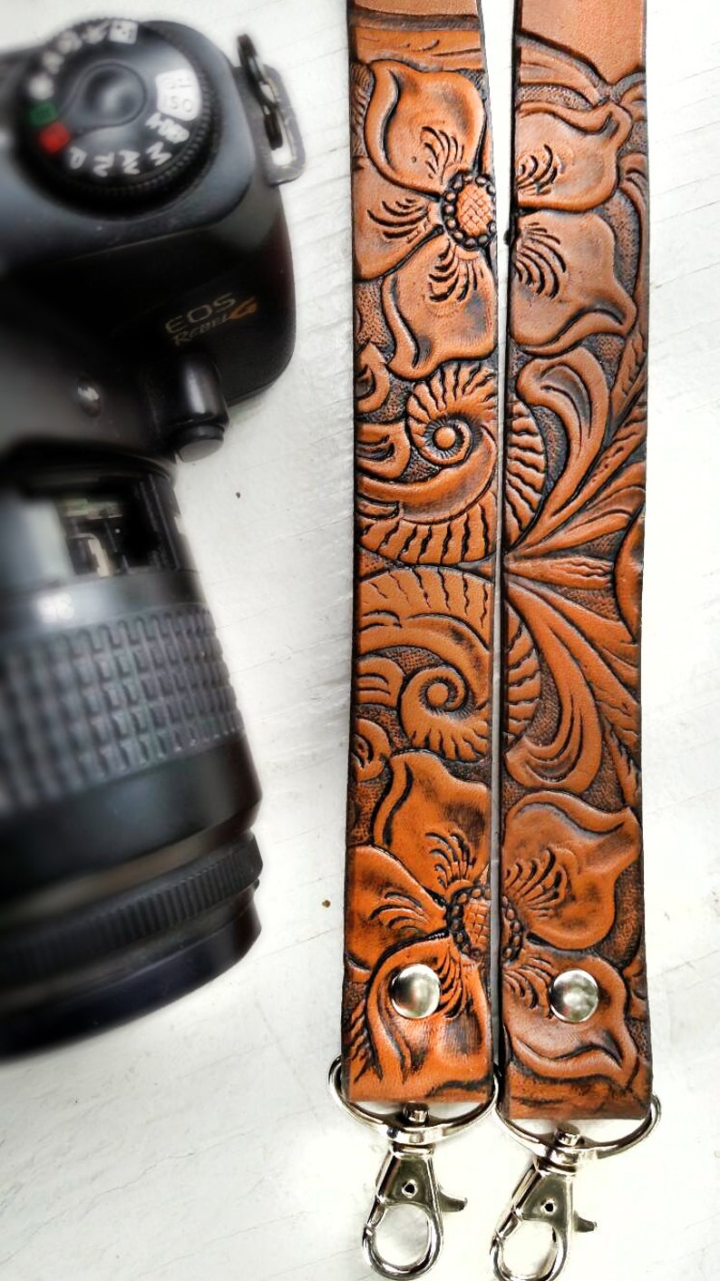 May include: A brown leather camera strap with a floral design. The strap has two metal clasps.