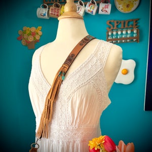 May include: A white crocheted dress with a brown leather strap with turquoise beads and fringe. The strap is worn over the shoulder and the fringe hangs down the front of the dress.