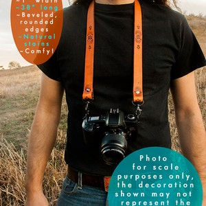 May include: A brown leather camera strap with decorative stitching. The strap is approximately 1 inch wide and 38 inches long. The strap is shown being worn by a person with long hair. The text "Photo for scale purposes only, the decoration shown may not represent the actual design of your strap choice" is visible in the image.