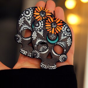 May include: A pair of black skull-shaped earrings with orange and white floral designs. The earrings are painted with white and turquoise details.