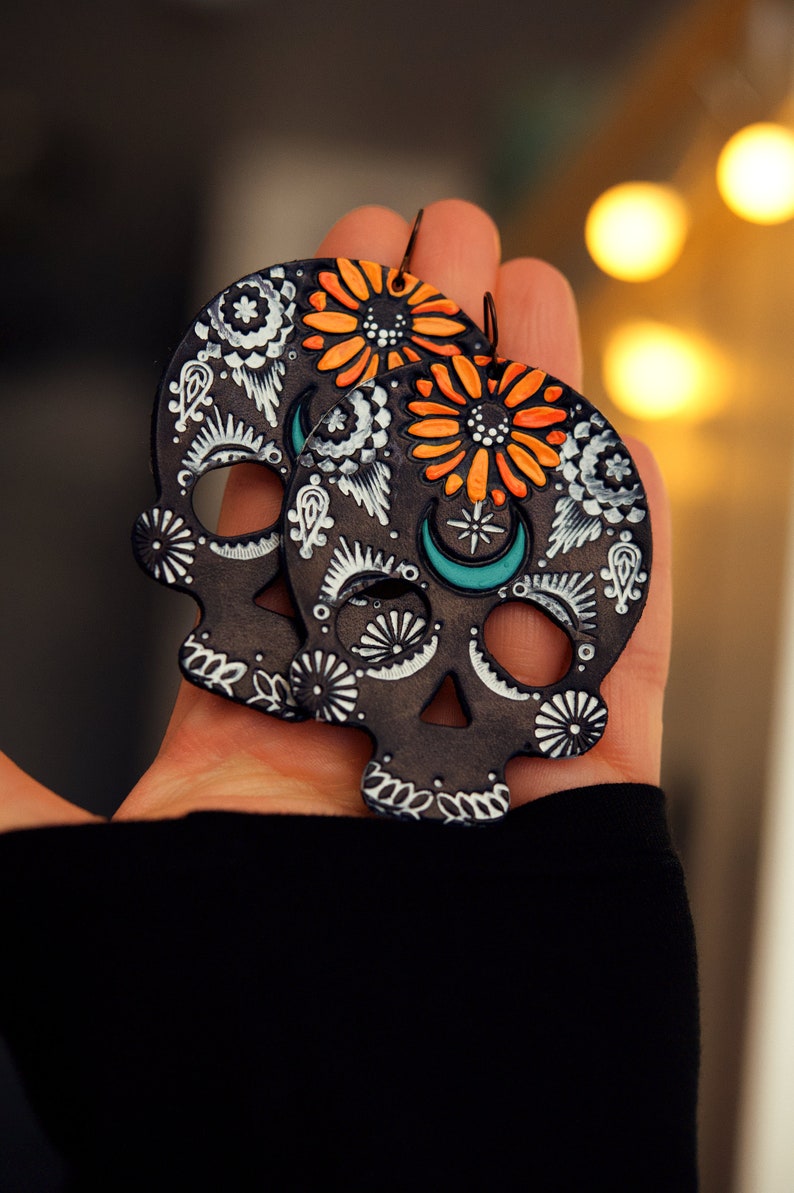 May include: A pair of black skull-shaped earrings with orange and white floral designs. The earrings are painted with white and turquoise details.