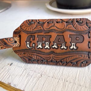 Peut inclure: Étiquette de bagage en cuir marron avec le mot "CHAP" en lettres blanches. L'étiquette présente des motifs floraux et de volutes en relief. Une boucle et une sangle argentées sont attachées à l'étiquette.