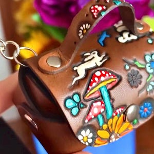 May include: A small brown leather purse with a handle, decorated with colorful painted mushrooms, flowers, and a white rabbit. The purse has a metal clasp and a key ring.