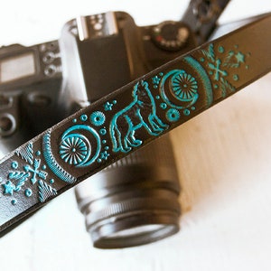 May include: A black leather camera strap with a teal blue wolf design and moon and star accents.