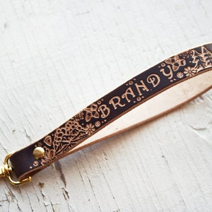 Könnte beinhalten: Brauner Leder-Schlüsselanhänger mit einem goldenen Metallverschluss. Das Leder ist mit einem floralen Muster und dem Text "BRANDY" geprägt.