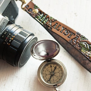 Peut inclure: Un bracelet de caméra en cuir marron avec le nom "Alysta" gravé dessus. Le bracelet est décoré de motifs floraux verts et bruns. Une boussole argentée et un appareil photo argenté sont également présents sur l'image.