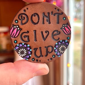 May include: A brown leather round pin with the text "DON'T GIVE UP" in black lettering. The pin is decorated with pink, white, and purple flowers.