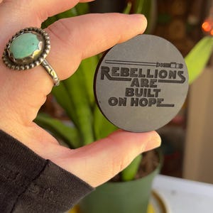 May include: A dark gray, circular coaster with the text "Rebellions Are Built On Hope" etched into the surface. The coaster is held in a hand, and a ring with a turquoise stone is visible on a finger.