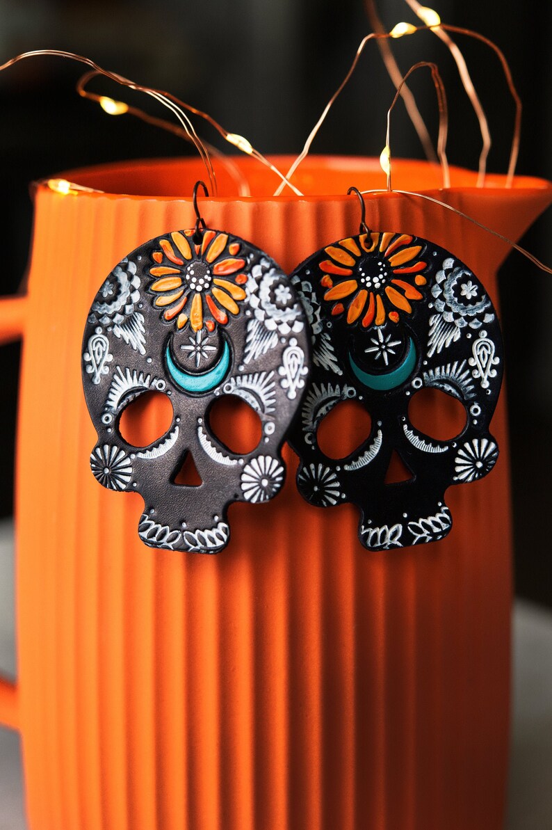 May include: A pair of black sugar skull earrings with white, orange, and turquoise details. The earrings are hanging from a light orange pitcher.