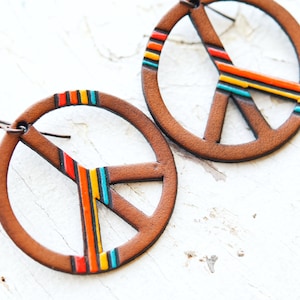 May include: A pair of brown leather earrings with a peace sign design. The peace sign is outlined in black and has a multi-colored stripe design in red, orange, yellow, turquoise, and blue.
