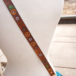 May include: A brown leather strap with a floral design. The strap features orange, white, green, and blue flowers painted on it. The strap is attached to a gold metal clasp.