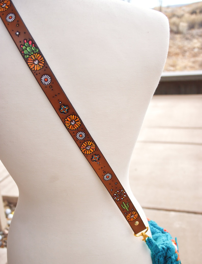May include: A brown leather strap with a floral design. The strap features orange, white, green, and blue flowers painted on it. The strap is attached to a gold metal clasp.