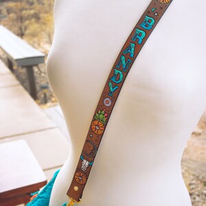 May include: A brown leather bag strap with the name "BRANDY" written in blue and green lettering. The strap is decorated with colourful flowers, cacti, and a skull design.