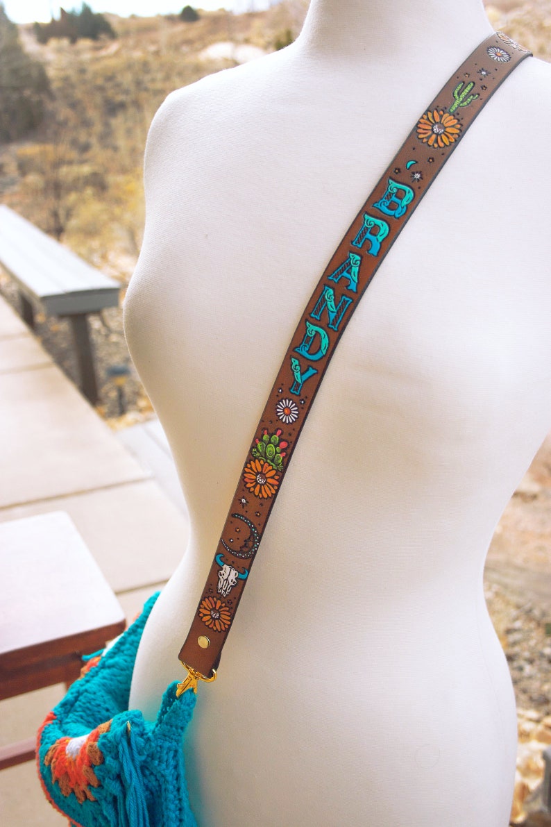 May include: A brown leather bag strap with the name "BRANDY" written in blue and green lettering. The strap is decorated with colourful flowers, cacti, and a skull design.