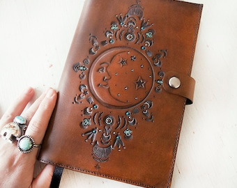 Pode incluir: Um diário de couro marrom com um design estampado de uma lua crescente e estrelas. O design é acentuado com pedras preciosas turquesa.