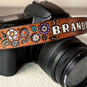 May include: A brown leather camera strap with a floral design and the name "BRANDY" embossed in white lettering.