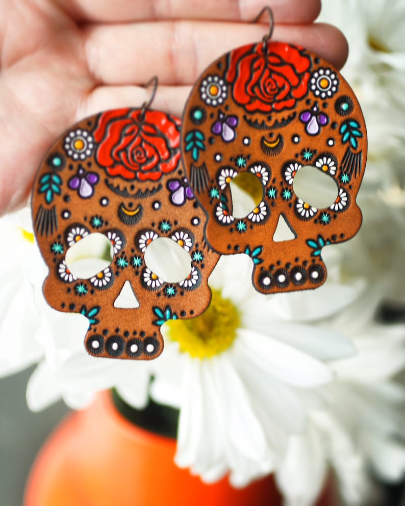 May include: A pair of brown leather earrings shaped like sugar skulls, decorated with colorful flowers, dots, and swirls. The earrings have a white background with turquoise, purple, and orange accents.