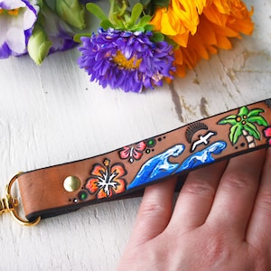 May include: A brown leather keychain with a colourful hand-painted design featuring palm trees, waves, a bird, and flowers. The keychain has a gold metal clasp and the text "Y" is visible.