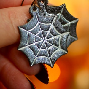 May include: A pair of silver glitter spiderweb earrings with a black background. The earrings are hanging from a person's hand.