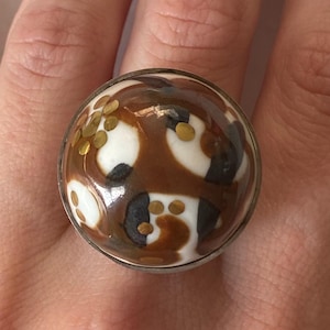 May include: A round ring with a large, patterned stone. The stone features a mix of brown, white, and black shapes, with small gold accents. The ring has a silver-colored band.