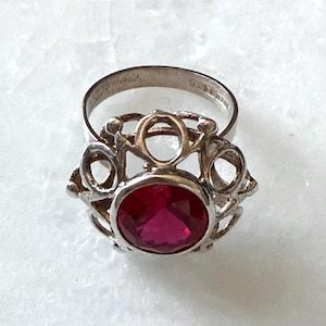 Könnte beinhalten: Ein silberner Ring mit einem ovalen, tiefroten Edelstein. Der Ring hat ein verziertes, durchbrochenes Design um den Edelstein herum. Das Band des Rings ist schlicht silberfarben. Der Ring ist vor weißem Hintergrund platziert.