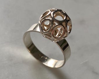 May include: A silver ring with a spherical cage design. The ring's band is polished silver, and the sphere is made of interconnected circular and star-shaped cutouts. The ring is displayed on a light-coloured surface.