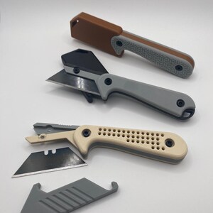 Könnte beinhalten: Drei Cuttermesser in verschiedenen Farben: Braun, Grau und Beige. Jedes Messer hat eine Klinge und einen Griff mit strukturierter Oberfläche. Ein Messer hat eine Klingenabdeckung. Eine Ersatzklinge und Klingenabdeckung sind ebenfalls sichtbar.