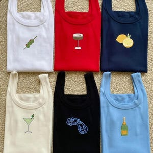 Puede incluir: Seis camisetas sin mangas en varios colores: blanco, rojo, azul marino, crema, negro y azul claro. Cada camiseta tiene un diseño bordado diferente de cóctel o comida, incluyendo aceitunas, un martini, limones, ostras y champán.