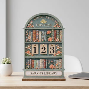 May include: A decorative bookshelf with the text "BOOKS READ THIS YEAR" and "SARAH'S LIBRARY". The shelf is painted in a muted green and adorned with floral accents and miniature books. It features numbered tags.