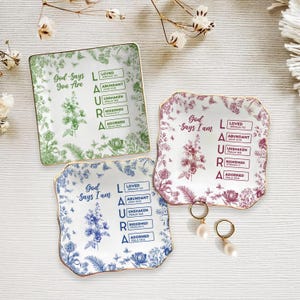 Puede incluir: Tres platos decorativos de cerámica con diseños florales en verde, burdeos y azul. Cada plato tiene un borde dorado y las palabras "Dios dice que eres" o "Dios dice que soy" con palabras inspiradoras. Pendientes de perlas también están en la imagen.