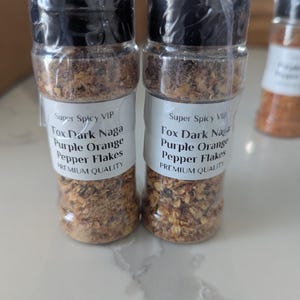 May include: Two small glass jars filled with dried orange and red pepper flakes. The labels on the jars read "Super Spicy VIP Fox Dark Naga Purple Orange Pepper Flakes Premium Quality".