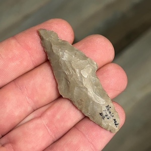 May include: A light gray, chipped stone arrowhead with a pointed tip and a slightly curved shape. The arrowhead has a rough texture and appears to be hand-crafted. Small text is visible on the arrowhead.