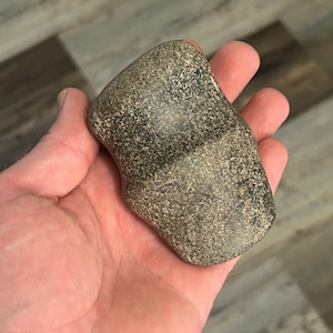 Puede incluir: Una mano sostiene una herramienta de piedra gris y negra. La piedra tiene una forma redondeada con una ranura en el medio. La superficie de la piedra está texturizada con pequeños puntos oscuros. La herramienta parece ser un artefacto antiguo.