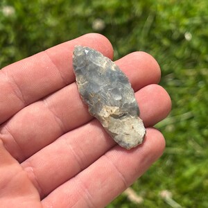 May include: A gray and white arrowhead, likely made of flint or chert, is held in a hand against a backdrop of green grass. The arrowhead has a pointed tip and a rough, textured surface, suggesting it is an ancient artifact.